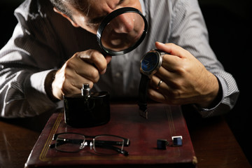 Mature man working at home as a watchmaker and jeweler composing an old clock using a screwdriver and a magnifying glass to be able to see closely