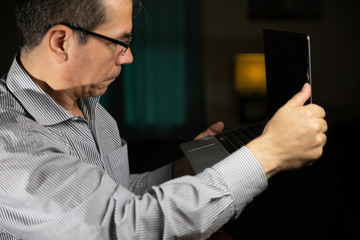 Mature age man with glasses at home working with his laptop sitting in the wooden dining room and using his mobile phone