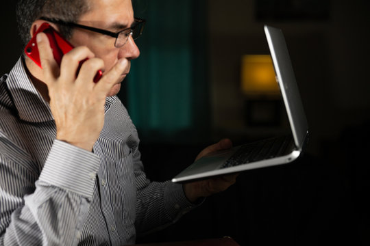Mature age man with glasses at home working with his laptop sitting in the wooden dining room and using his mobile phone