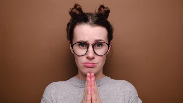 Pleading female in glasses looks gloomy and desperate, beggs for forgiveness feels sorry and guilty, keeps palms together, standing over brown background. Cute adorable girl pleads about something
