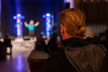 Closeup of male with blonde ponytail listening to speech delivered by speaker in illuminated auditorium hall in world and spoken word festival