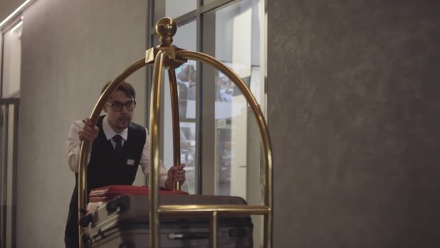 Tracking shot of male bellhop in uniform and glasses pushing luggage cart in hotel
