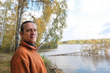 A man in an orange sweater against the background of a river bank with a birch grove. Autumn forest