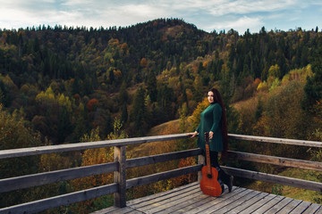 Fototapeta na wymiar Long-haired girl holds guitar, standing near parapet in mountains in autumn.