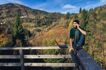 Fototapeta na wymiar bearded businessman sits on railing, works on laptop and talks on phone. Freelancer hipster on vacation in mountains.