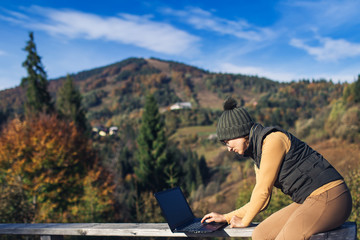 businesswoman sitting on railing and working on laptop. Freelancer hipster on vacation in mountains.
