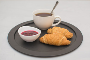 Morning coffee in a white cup with croissants and raspberry jam on a black tray.