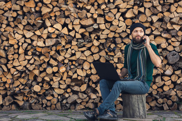 bearded freelancer is working on laptop and talking on phone against background of wall of firewood.