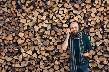bearded man in bluetooth headphones listening to music outdoors near pile of firewood.