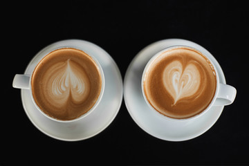 Two white cups of Cappuccino coffee with heart-shaped milk foam. Top view on two cups of latte coffee with heart figure on milk foam isolated on black background. Concept of breakfast. View from above
