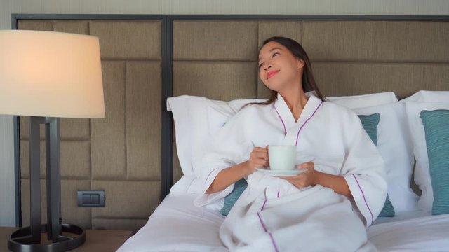 A young Asian woman in white bath robe enjoys coffee in bed. Medium shot.