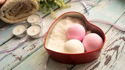 Aromatic balls and bath salt in a heart-shaped box, next to candles, flowers and a towel on a wooden background. Items for spa treatments.