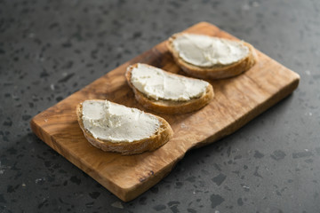 Three ciabatta slices with cream cheese on olive wood board on terrazzo surface