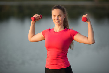 beautiful young woman in sportswear is engaged in summer training in a park near the lake with dumbbells in her hands © Alexandr