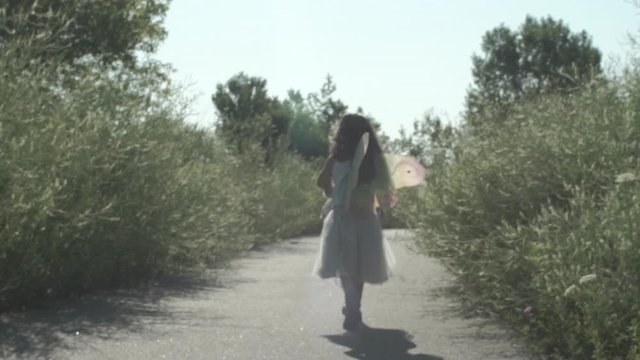 Child girl walks on flower path in sun wearing fairy wings wide frame child walks away from camera as camera follows in slow motion