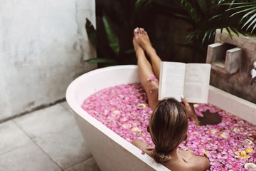 Woman reading book while relaxing in bath tub with flower petals