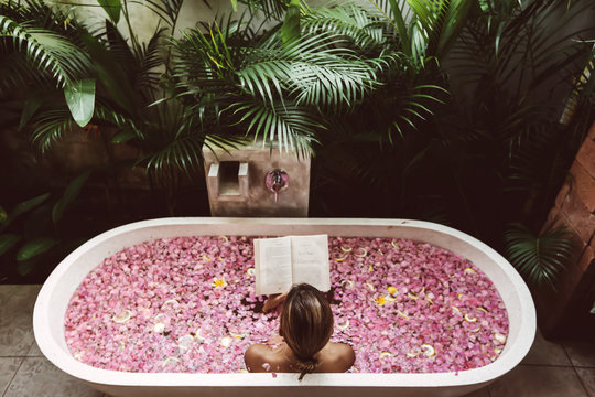 Woman reading book while relaxing in bath tub with flower petals