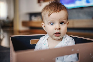 Confused little blond boy with blue eyes sitting in box and looking at camera with mouth opened....