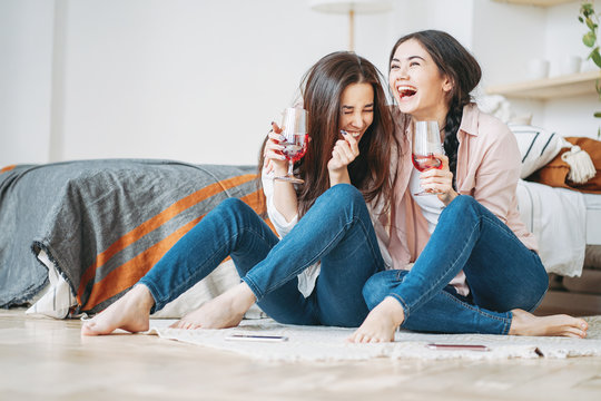 Young carefree laughing brunette girls friends in casual with glasses of wine having fun together on home party