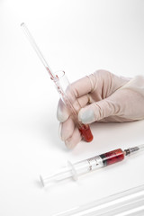 the gloved hand of a laboratory assistant holds a test tube with a blood sample on a white background. also in the photo is a syringe with a blood sample