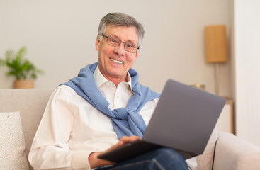 Cheerful Elderly Man With Laptop Computer Sitting On Sofa Indoor
