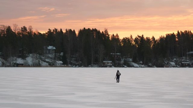 Fisherman walking on frozen lake searching for the perfect spot for ice fishing, golden morning sky