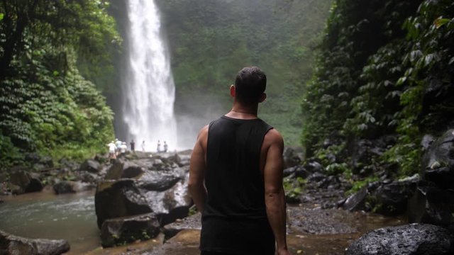 A young, fit man is walking by himself towards a waterfall in the depth of the Bali jungle. Holding a big and professional DSLR camera to take photos and videos. Slow motion.