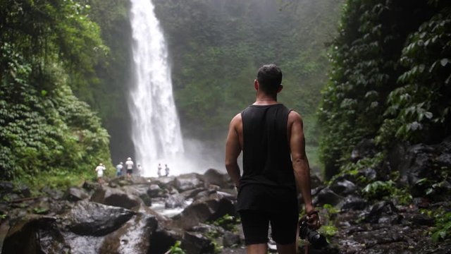 A young, fit and muscular man is walking by himself towards a waterfall in the depth of the Bali jungle. Holding a big and professional DSLR camera to take photos and videos. Slow motion.