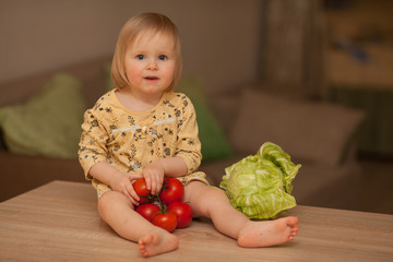 Fototapeta premium Cute little girl in yellow dress, vegetarian, eating vegetables cabbage and tomatoes in the kitchen. Concept: healthy food, diet, vegetarianism. Vegan food.