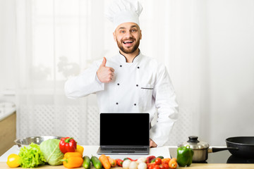 Chef Showing Laptop Blank Screen Gesturing Thumbs-Up Standing In Kitchen