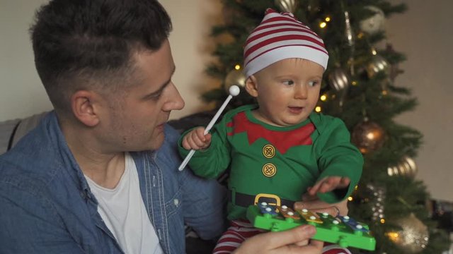 Dad and infant son wearing elf costume play with toy xylophone beside the Christmas tree. Baby boy playing with daddy during the holidays. Baby banging on xylophone with hand.