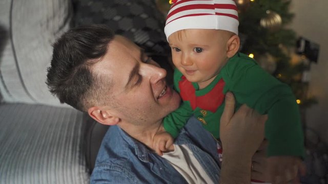 Smiling father playing with young child dressed on elf costume near Christmas tree with lights shining. Baby and dad bonding during holidays. Man holding son smiles with happiness.