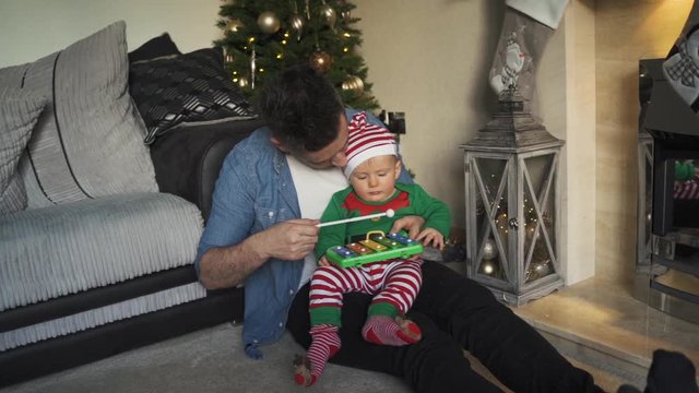 Slow motion view of dad and son playing with a toy xylophone at Christmas near decorated tree. Child in elf pajamas sitting on lap of father. Parenthood and holiday concept.