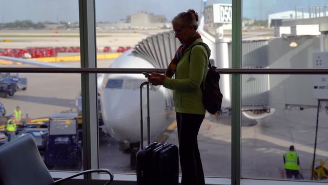Silhouette of woman with luggage checking her cellphone in front of airplane getting ready to board.