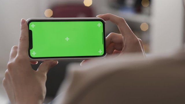 Girl using smartphone with green screen in living room watching movie, video content. Young woman at home lying on couch. Tracking markers. Smart phone in a horizontal position. Indoors. Closeup