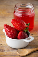Close-up of white bowl with strawberries, wooden spoon and recycled glass jar with strawberry jelly, on rustic wooden table, in vertical