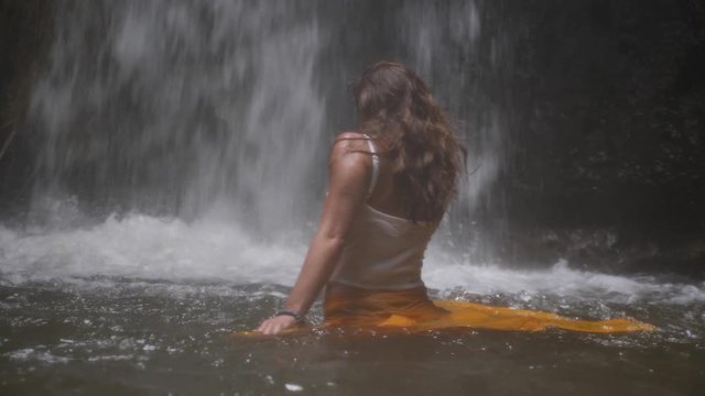 A young woman with long, dark hair is spinning and dancing around in a very sensual way in slow motion with a waterfall in the background in the jungle of Bali.