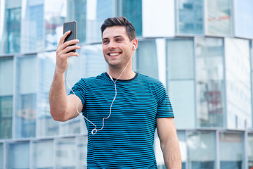 smiling young man holding mobile phone taking selfie outside in city