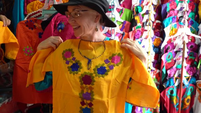 Mature woman holds up a yellow huipil blouse and then smiles looking at herself in Merida, Mexico.