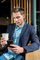 young businessman sitting at city cafe with cup of coffee and looking at mobile phone