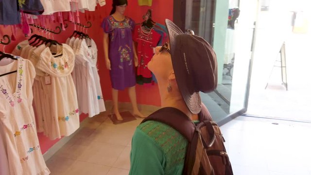 Camera follows mature woman looking at beautiful display of women’s huipil blouses in Merida Mexico store.