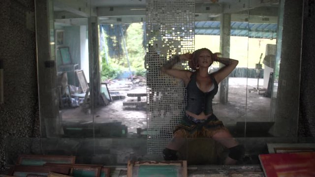 A young woman with a cool jeans vest, skirt and boots outfit is dancing sensually against a mirror glass wall with pieces of mosaic in a dark, abandoned room.