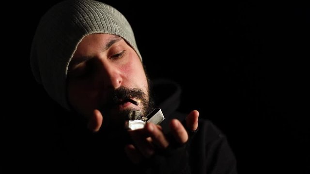 Close up of a contrasted portrait of young man with beard and hat lighting cigarette and looking to camera
