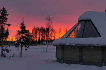 Cottage with large windows in snow landscape with sunset and orange sky in background, Lapland, Finland