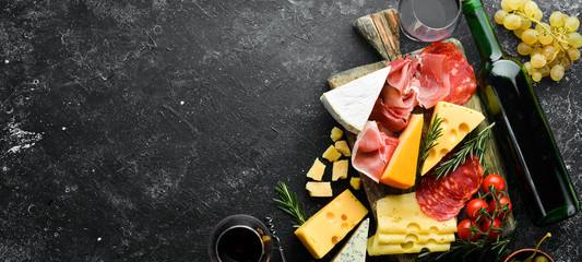 Obraz na płótnie Canvas Appetizers table with italian antipasti snacks and wine in glasses. Cheese, wine, salami and prosciutto on a black stone background. Top view. Free space for your text.