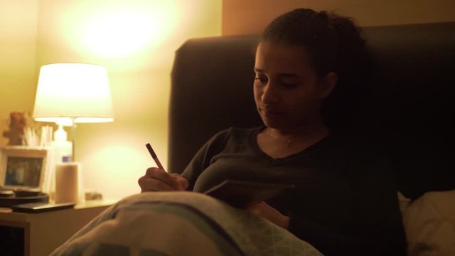 Girl is lying in bed writing in diary in the light of a bedside lamp.