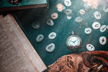 Astrology and esotericism. On the dark surface lie a watch on a chain, fortune-telling Runestones, a book, and a cloth. The view from the top. Dust and light