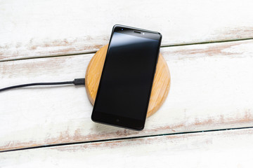 Concept of modern technologies. The black smartphone is on a wireless charger. White wooden background with diagonal stripes. Copy space. Flat line