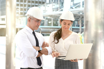Two engineer wearing white safety helmet using a laptop together, male and female discussing about project work while walking on the way outside office in downtown city