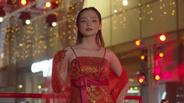 Beautiful Asian woman dressed to celebrate the Chinese New Year poses in her red sleeveless dress with the bokeh of the night lihts in the background